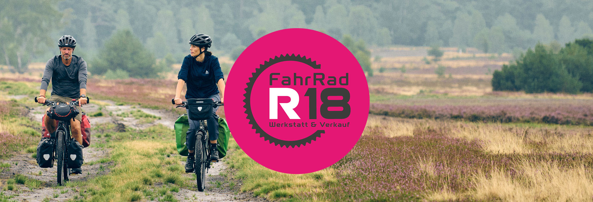 Das Logo des münchner Fahrradladens FahrRad R18 ist zu sehen. Im Hintergrund sieht man zwei Radfahrende auf ihren Bikepacking-Bikes. Das Logo ist ein graues Kettenblatt mit der Aufschrift "FahrRad R18 - Werkstatt und Verkauf", vor einem pinken Kreis.