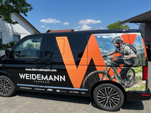 Ein schwarzer Van mit Fahrzeugbeklebung: Ein Mountainbiker und ein großes, oragenes W sind zu sehen. Außerdem das Logo der Firma Weidemann Fahrrad aus Überlingen. Im Hintergrund sind ein Baum und ein Haus zu sehen. (Seitenansicht des Vans)