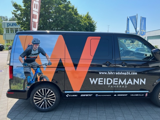 Ein schwarzer Van mit Fahrzeugbeklebung: Ein Mountainbiker und ein großes, oragenes W sind zu sehen. Außerdem das Logo der Firma Weidemann Fahrrad aus Überlingen. Im Hintergrund sind ein Baum und ein Haus zu sehen.