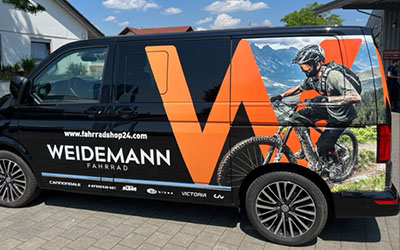 Ein schwarzer Van mit Fahrzeugbeklebung: Ein Mountainbiker und ein großes, oragenes W sind zu sehen. Außerdem das Logo der Firma Weidemann Fahrrad aus Überlingen. Im Hintergrund sind ein Baum und ein Haus zu sehen.
